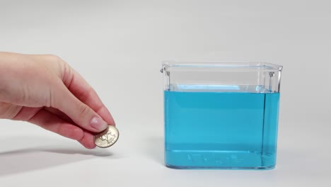 a hand drops a metal coin into a clear container filled with blue liquid, observing its sinking behavior