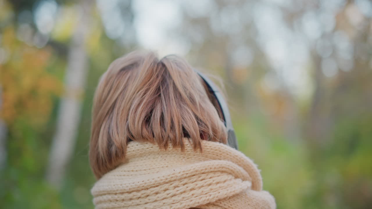 back view of middle aged woman wearing headphones strolling down sunlit autumn forest path with blurred background showcasing cozy scarf and bob haircut while enjoying peaceful walk in nature
