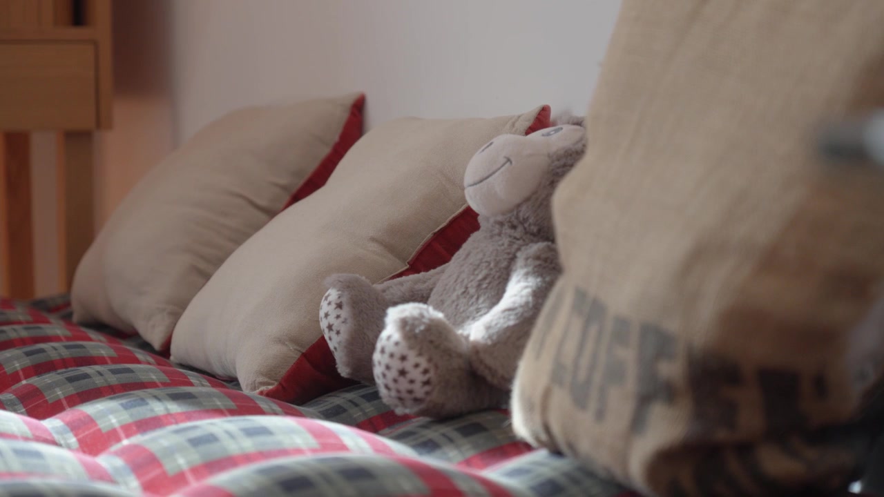 Inquadratura ad arco lenta che rivela un morbido peluche appoggiato a cuscini su un letto a quadri rossi, luce diurna interna soffusa, atmosfera casalinga accogliente.