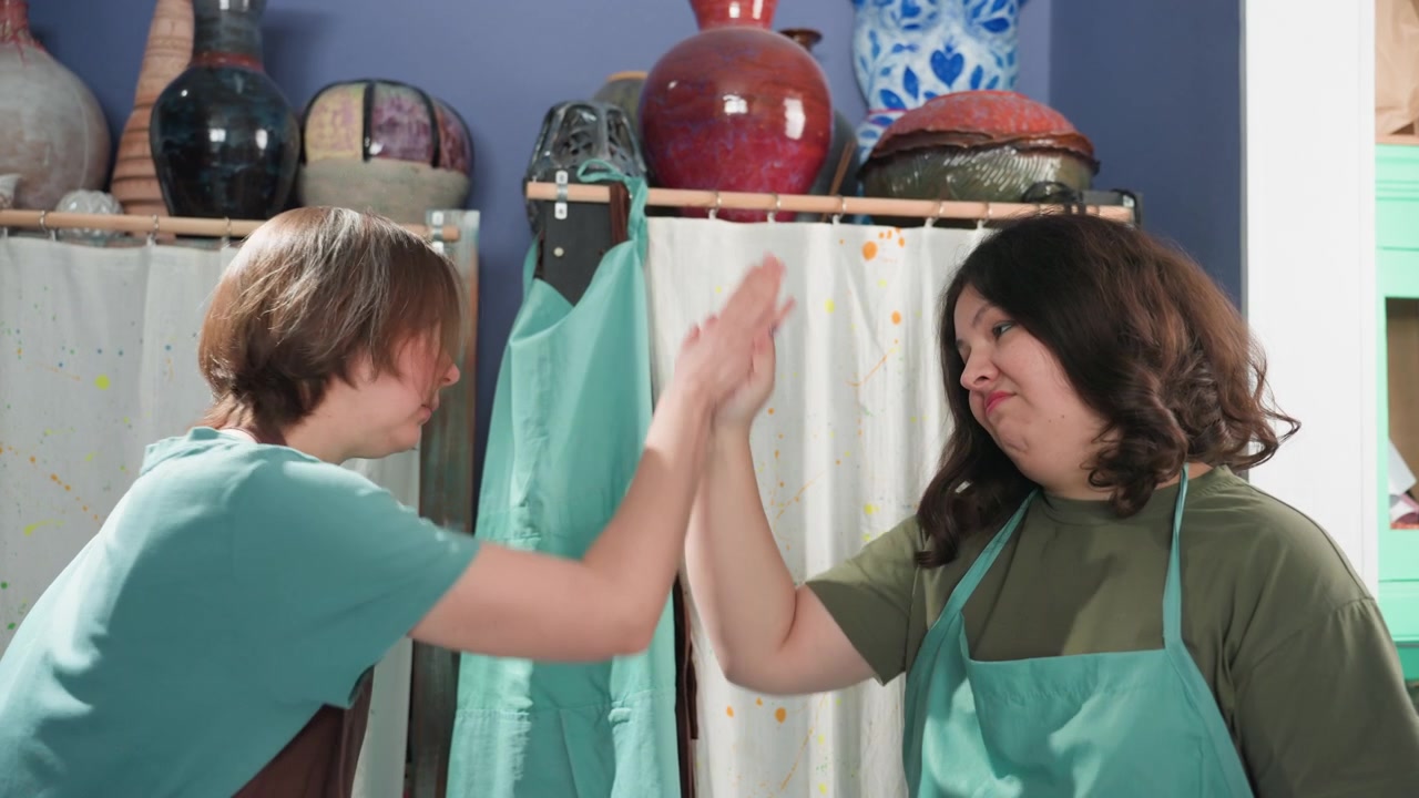 Due donne in uno studio di ceramica che condividono battute scherzose e sorrisi mentre si aggiustano i grembiuli, scaffali di smalti e vasi di ceramica dietro di loro, scambio amichevole e un momento di "cinque" casuale