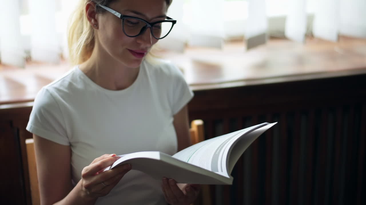Blonde woman wearing black reading glasses and a white T-shirt, reading a book on a chair in front of a window