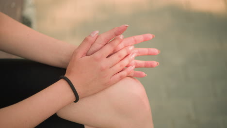 vista parcial de una dama sentada en un banco de madera al aire libre con las piernas cruzadas, abrazando y frotando suavemente las manos juntas descansando en la rodilla, usando un brazalete de mano negro