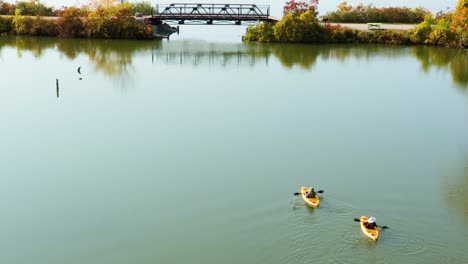 Una-Vista-Aérea-De-Dos-Personas-Navegando-Lentamente-En-Kayak-Alejándose-De-La-Cámara-Hacia-Un-Puente-Peatonal