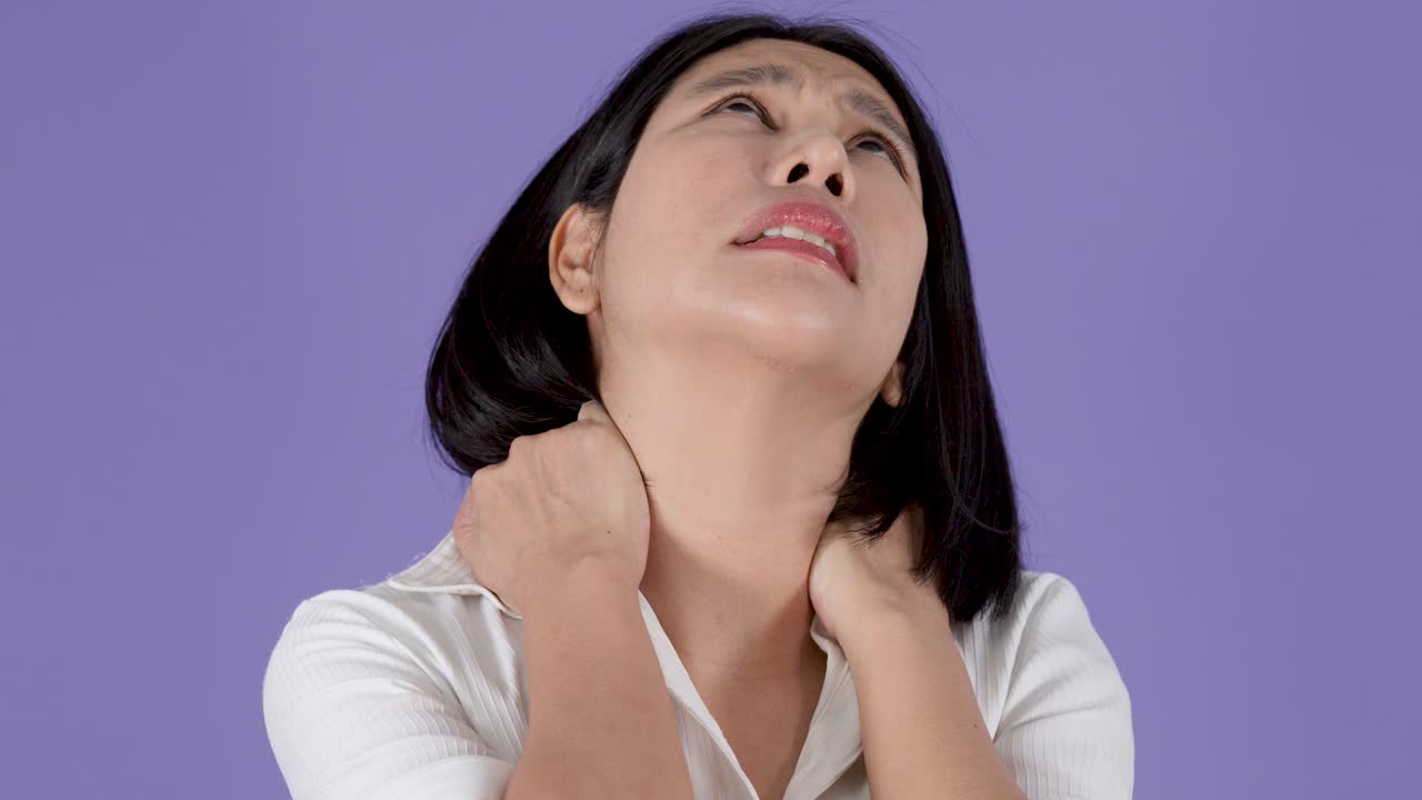 Donna asiatica si massaggia collo e spalle indolenziti su sfondo viola, esprimendo dolore muscolare legato al lavoro in ufficio