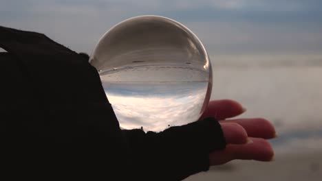 movimiento lento de una mano femenina con un guante sin dedos sosteniendo una bola de cristal que refleja al revés una playa y un paisaje marítimo en un día nublado y ventoso de invierno