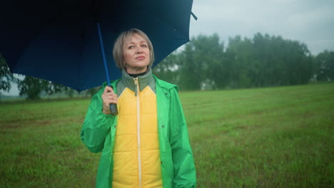 mujer en chaqueta amarilla y verde bajo impermeable verde de pie en un vasto campo de hierba sosteniendo un paraguas abierto, mira en la distancia con árboles en el fondo lejano bajo un cielo nublado