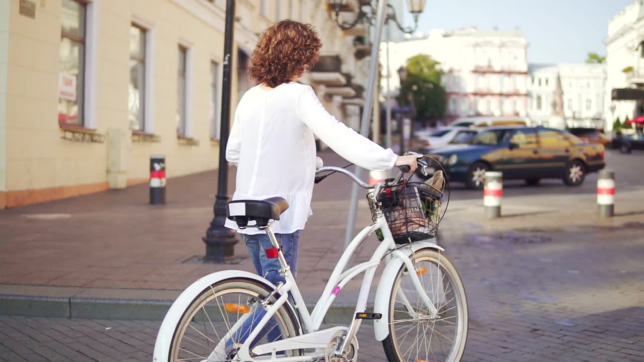 задний вид брюнетки в белой рубашке и голубых джинсах, идущей по городской улице, держащей ручку велосипеда с