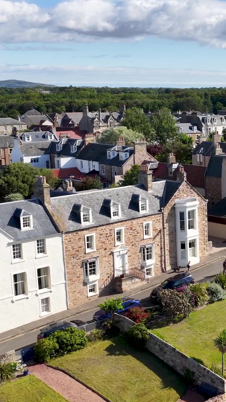 Un drone plane au-dessus de toits historiques ensoleillés, révélant un charmant paysage urbain côtier et un environnement verdoyant luxuriant.