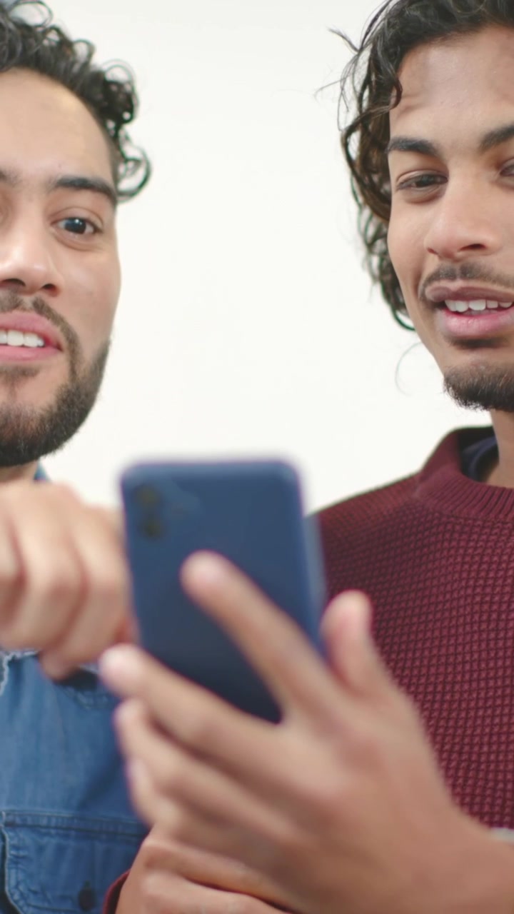 Vídeo vertical: Reagindo a um ping, dupla inclinada sobre telefone azul em ambiente interno, com suéter bordô e camisa jeans.