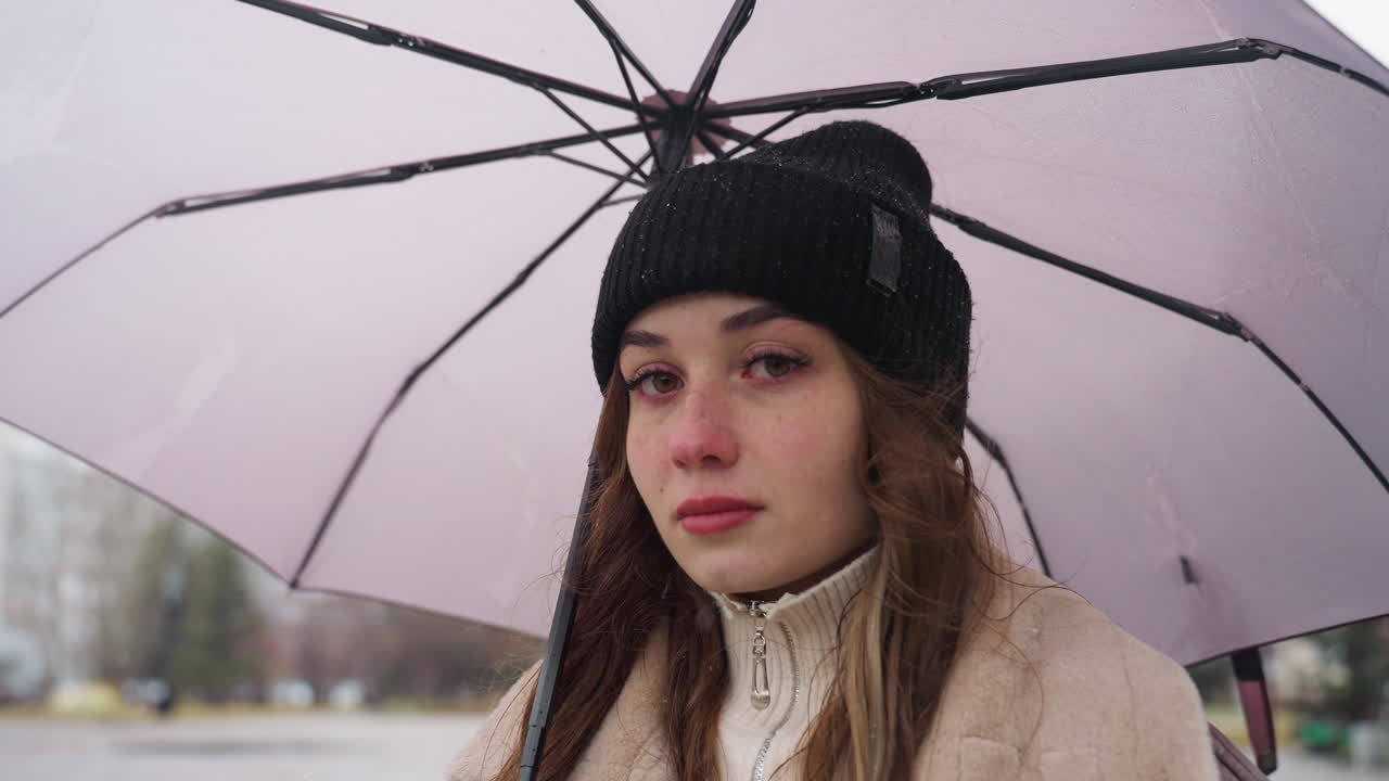 Young woman with serious expression standing still, holding umbrella, wearing black knit cap and brown shearling jacket, under cold overcast sky with light snowfall, contemplative mood
