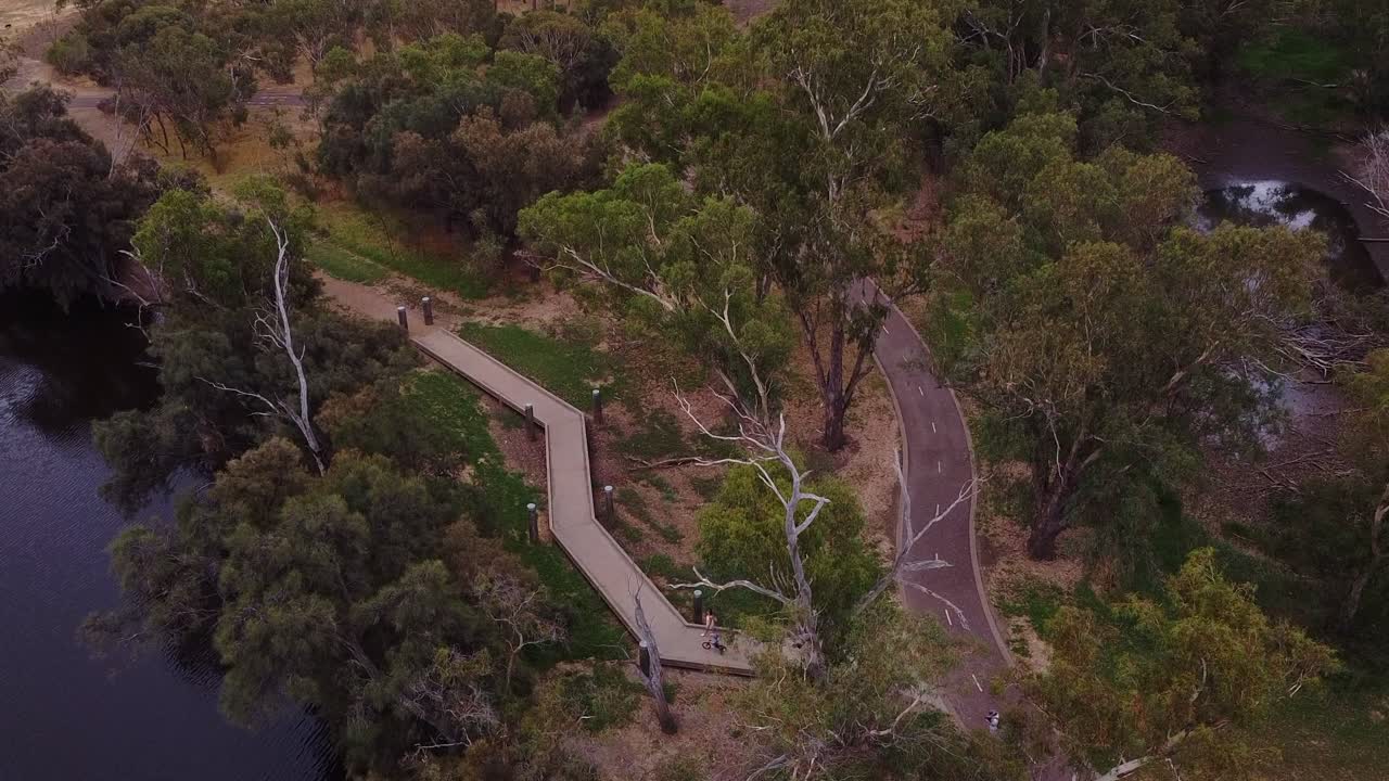 강변 의 자전거 도로 근처 의 보드워크 를 따라 타고 있는 아이 와 함께 어머니 를 바라보는 공중 전망