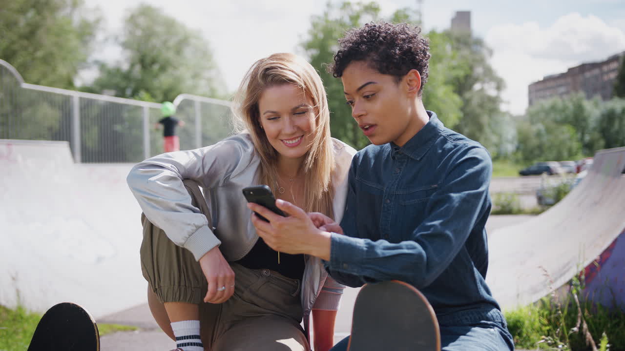 Two Female Friends Looking At Mobile Phone In Urban Skate Park And Laughing
