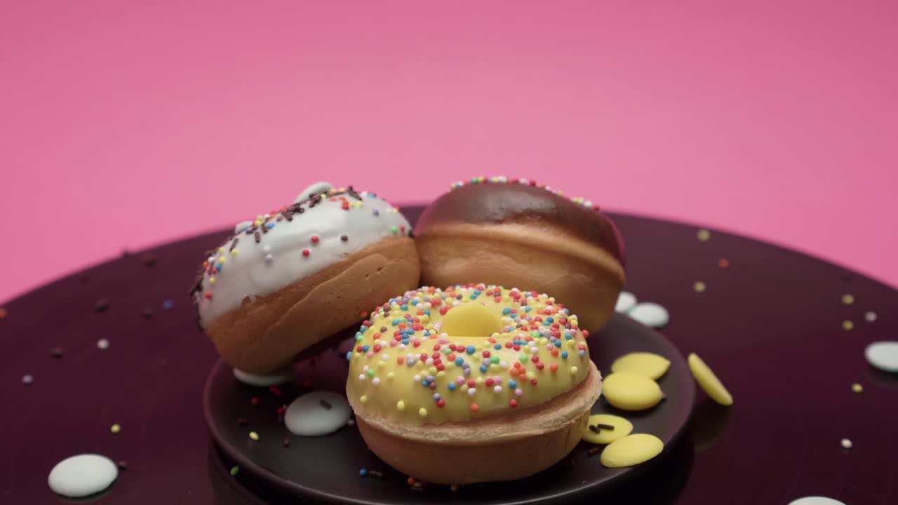 Three American donuts spin in circles on a black and pink surface. Topped with black, yellow and blue chocolate and choco and colorful crumbs