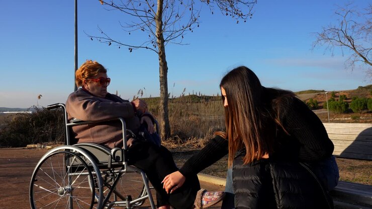 Old woman in wheelchair assisted by granddaughter outdoors on a sunny day