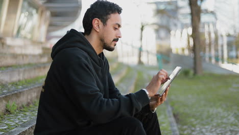 Side-view-of-young-Arabic-handsome-man-with-dark-curly-hair-and-beard-in-black-hoodie-sitting-on-stairs-outside