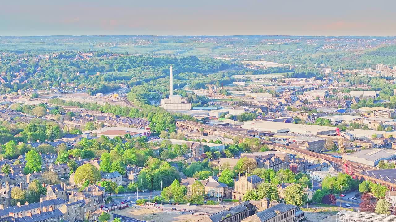 Wide aerial static shot of Huddersfield, England, capturing industrial buildings, the tall incinerator stack, rail tracks, and scattered tree canopies under soft afternoon light.