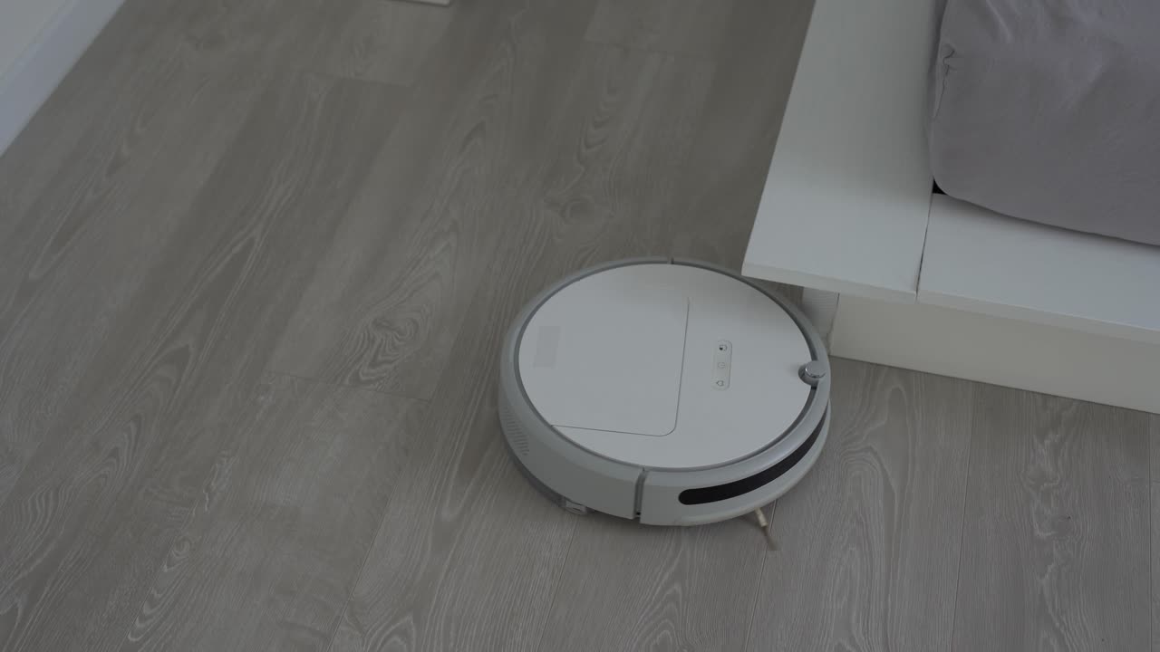 White robotic vacuum cleaner on laminate floor cleaning dust in living room interior. Smart housekeeping technology.