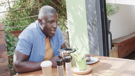 happy african american man talking on smartphone at table outside coffee shop, slow motion