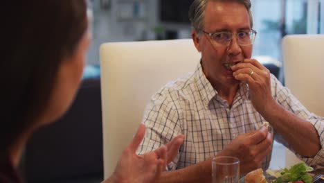 Smiling-caucasian-grandfather-talking-to-daughter-in-law-and-eating-at-table-during-family-meal