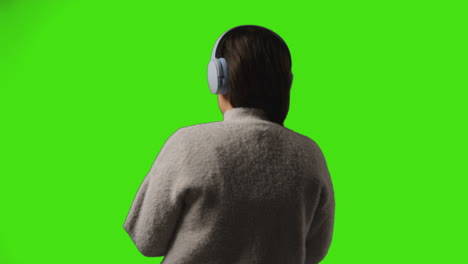 rear view of young woman wearing wireless headphones streaming music from mobile phone against studio green screen 1