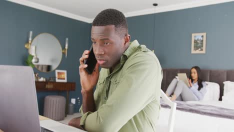 Video-of-diverse-couple-in-bedroom,-the-man-sitting-using-smartphone-and-laptop-in-foreground