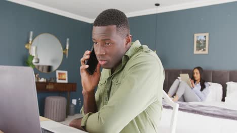 video of diverse couple in bedroom, the man sitting using smartphone and laptop in foreground