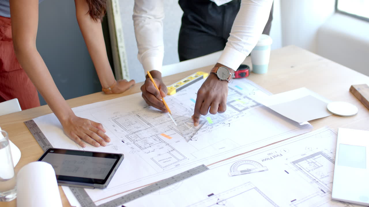 Discussing architectural plans, diverse colleagues pointing at blueprints on desk with tablet