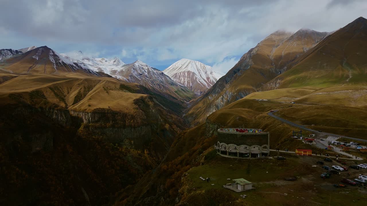Aerial panorama of the Georgian Friendship Monument with snow-capped Caucasus peaks and scenic winding mountain road