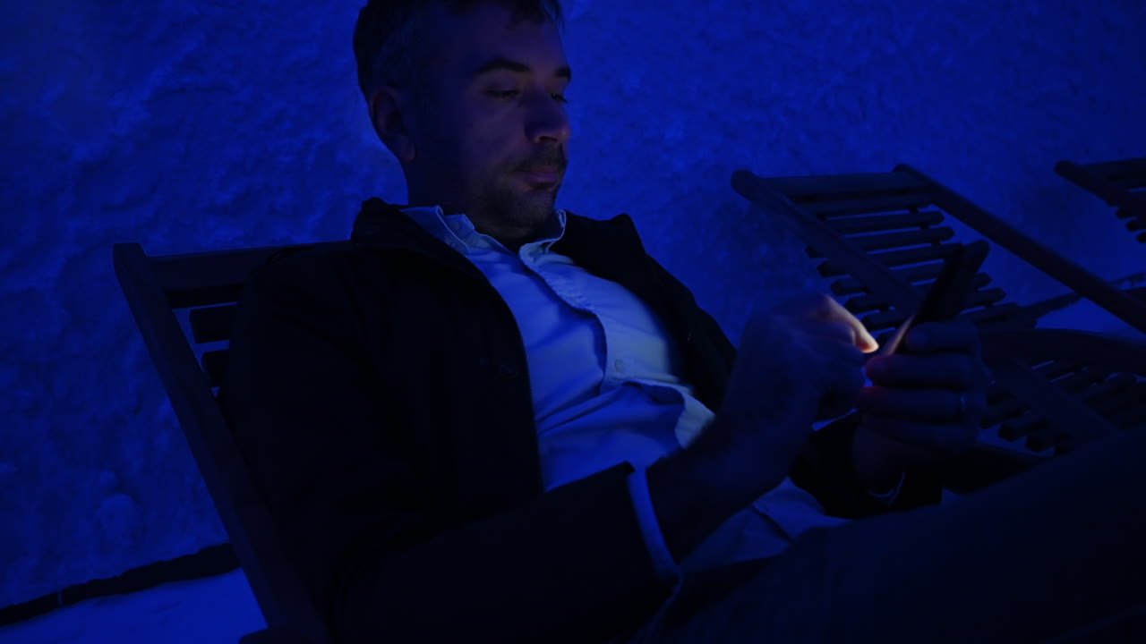 Man relaxing on a wooden chair in a salt room with blue light, working on mobile phone