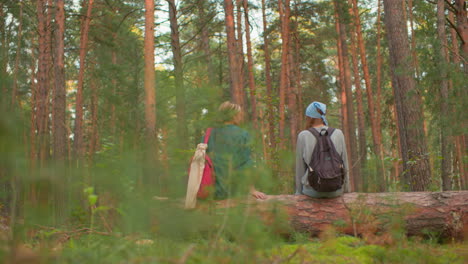 two hikers with backpacks rest on fallen tree in lush forest, one wearing blue bandana and gray sweater, other in green shirt with red bag, they appear relaxed, surrounded by tall trees and greenery