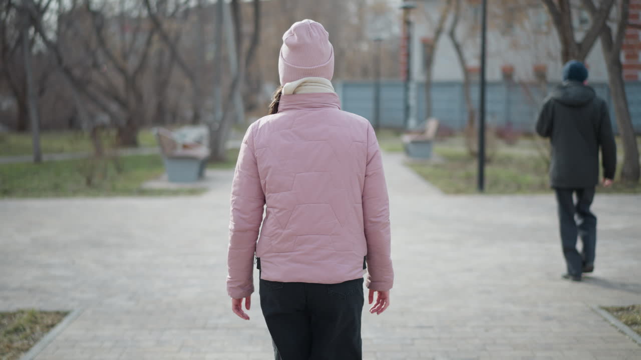Woman in pink jacket and hat walks along paved urban path in winter park as man walks ahead in same direction, surrounded by leafless trees, benches, and early spring grass in calm, quiet setting