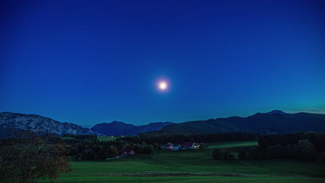 statische brede opname van een pittoresk landschap in oostenrijk 's nachts met intense blauwe hemel bij volle maan over de majestueuze bergketens en dichte bossen met weiden