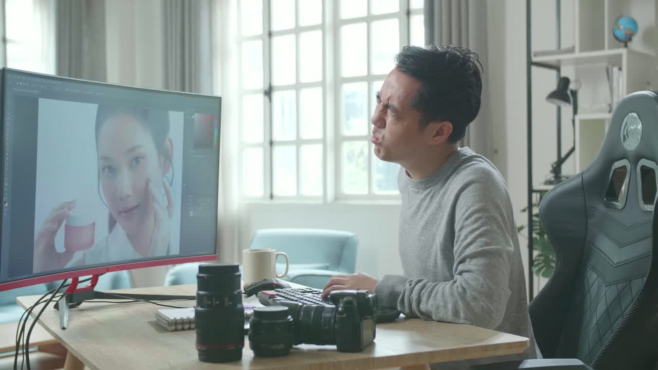 アジアのカメラマンが長袖のtシャツを着てデスクトップコンピュータを使って自宅で働いています
