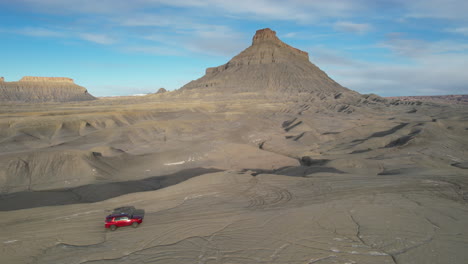 Vista-Aérea-Del-Vehículo-Todoterreno-Rojo-Que-Se-Mueve-Por-Un-Camino-De-Tierra-En-Un-Paisaje-Desértico-Bajo-Factory-Butte,-Utah,-EE.UU.