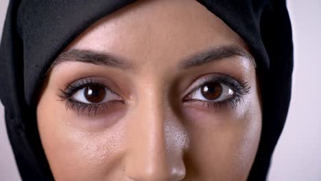 eyes of young serious muslim girl in hijab is watching at camera, blinking, grey background