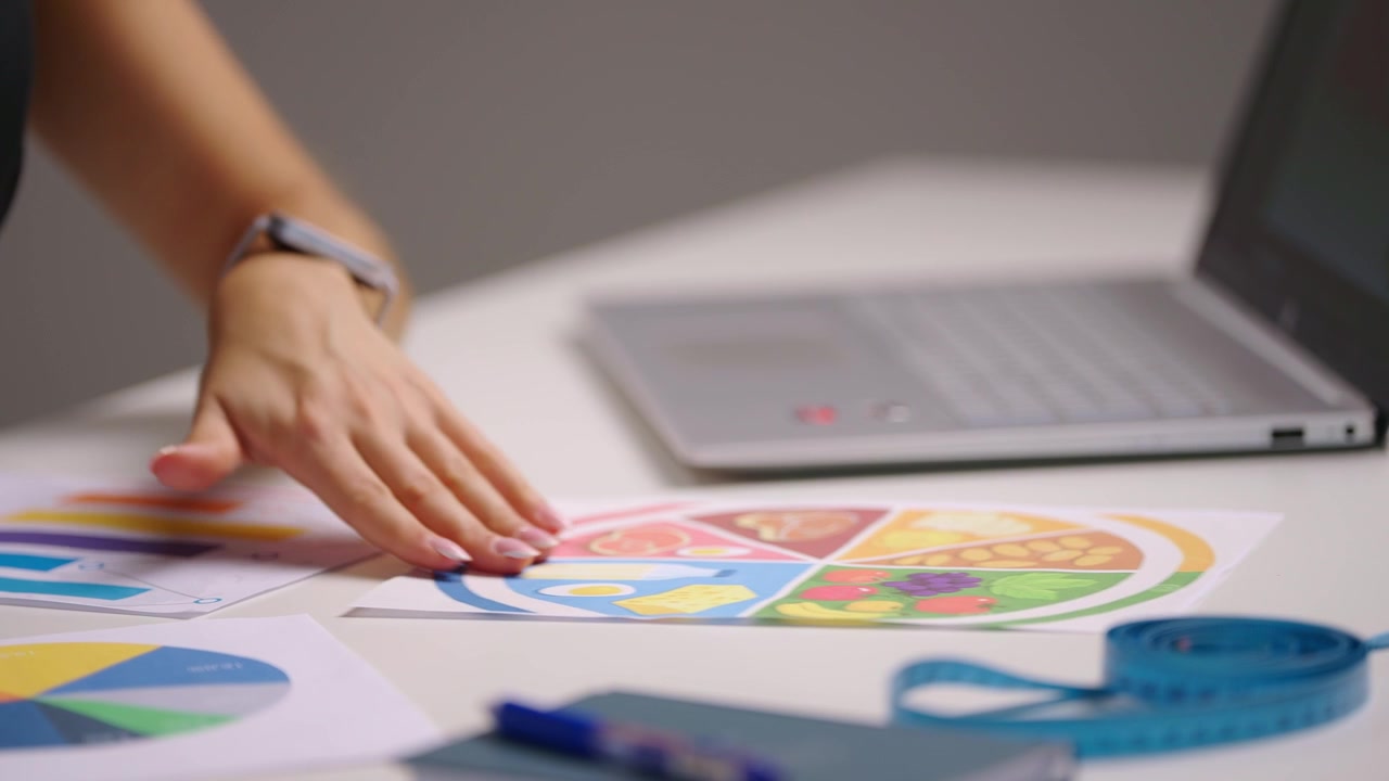 Vista laterale di una nutrizionista che lavora al laptop mentre esamina grafici e diagrammi dietetici colorati sulla scrivania, creando un piano nutrizionale personalizzato con analisi dei dati per obiettivi di stile di vita sano