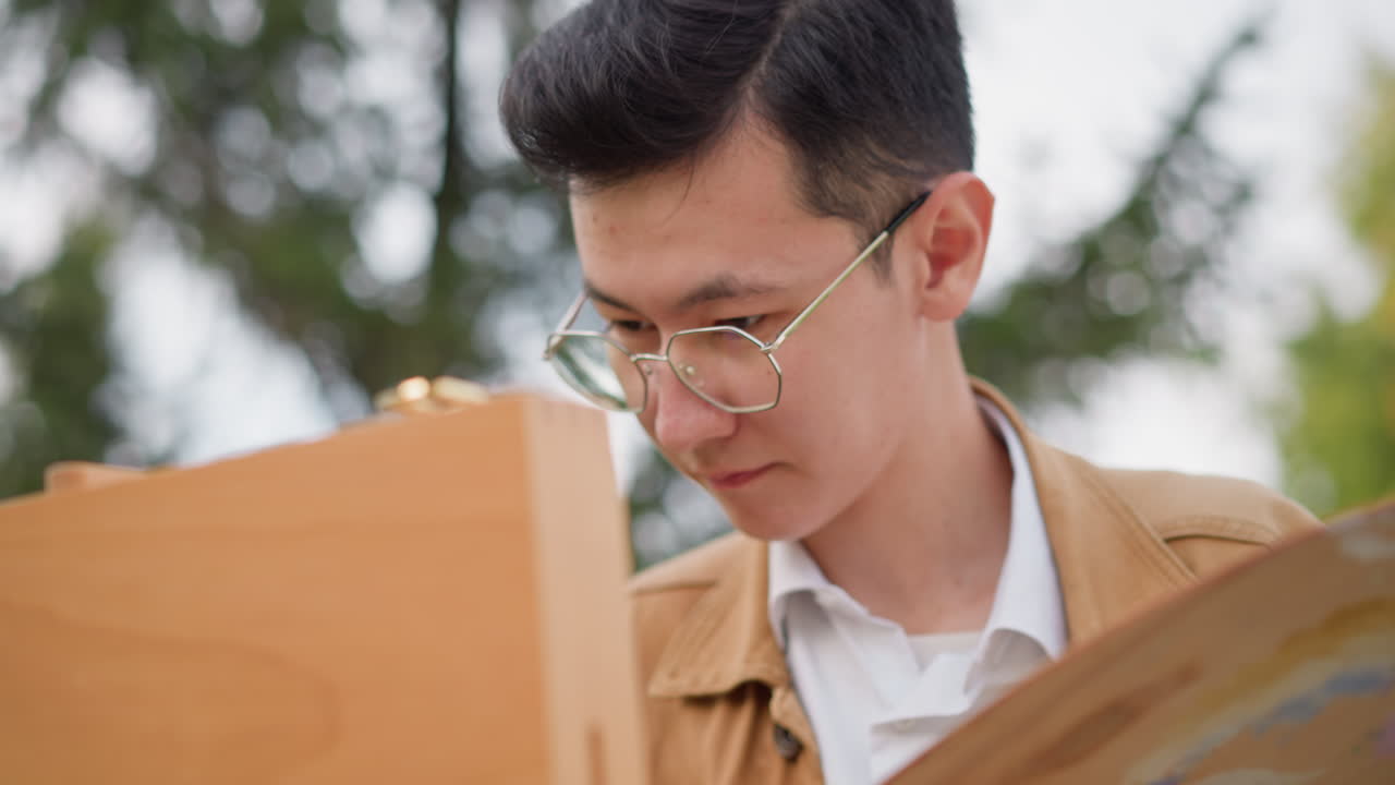 屋外のイーゼルで絵を描くアジア人男性。葉が生い茂る公園で、メガネと茶色のジャケットを着用し、真剣な表情で人物画の練習をしている。 キャンバスにスケッチをし、筆を調整し、コンポジション・リファレンスを調べている様子をクローズアップフレーミングで捉えています。