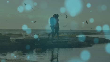 blue spots of light floating against african american couple walking on the rocks near the sea