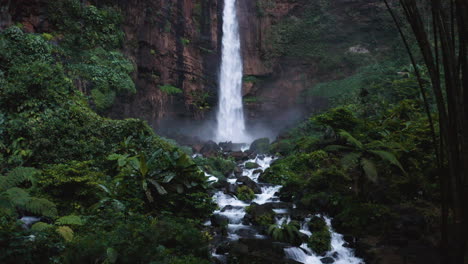 aerial view tilting slowly toward the air terjun kapas biru waterfall, in cloudy east java, indonesia