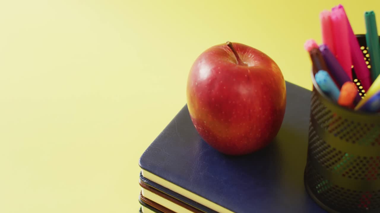 video de una pila de libros, marcadores en un contenedor y una manzana sobre un fondo amarillo