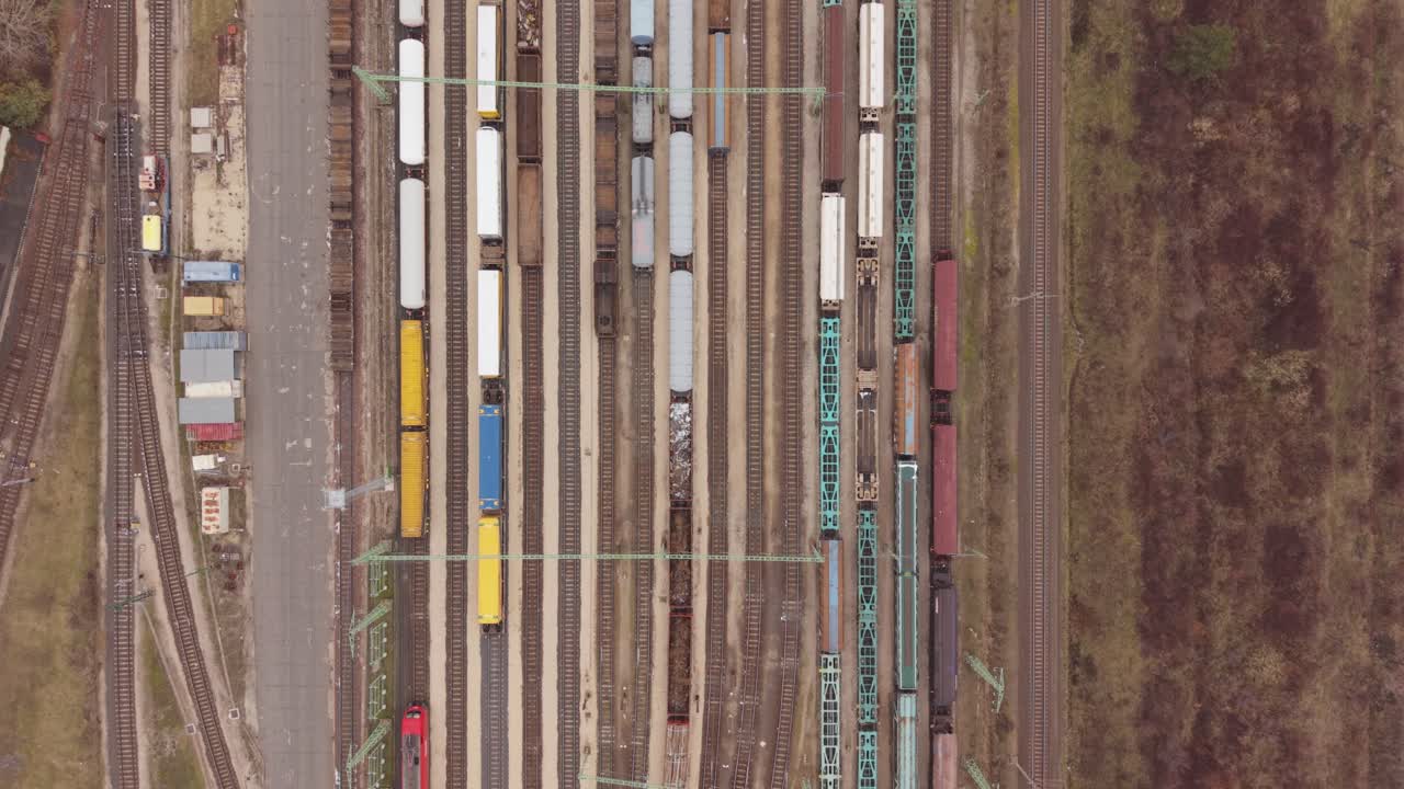 A straight-down aerial view of a busy freight railway yard filled with cargo wagons arranged on multiple parallel tracks. Drone follow a leaving train