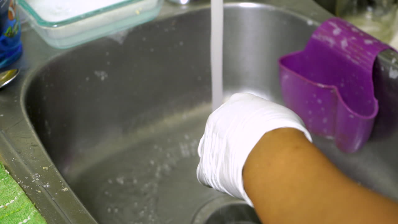 Person wearing gloves is doing household chores like washing dirty dishes under running water