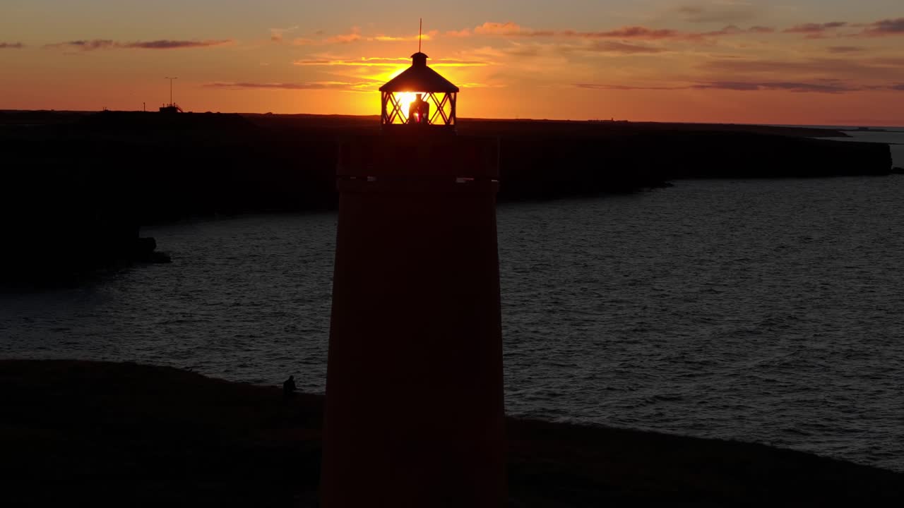 Um drone abrangente capta o vibrante farol amarelo Hólmsbergslviti contra um céu laranja ardente. Homem passeando em falésias escarpadas em Keflavik, Islândia