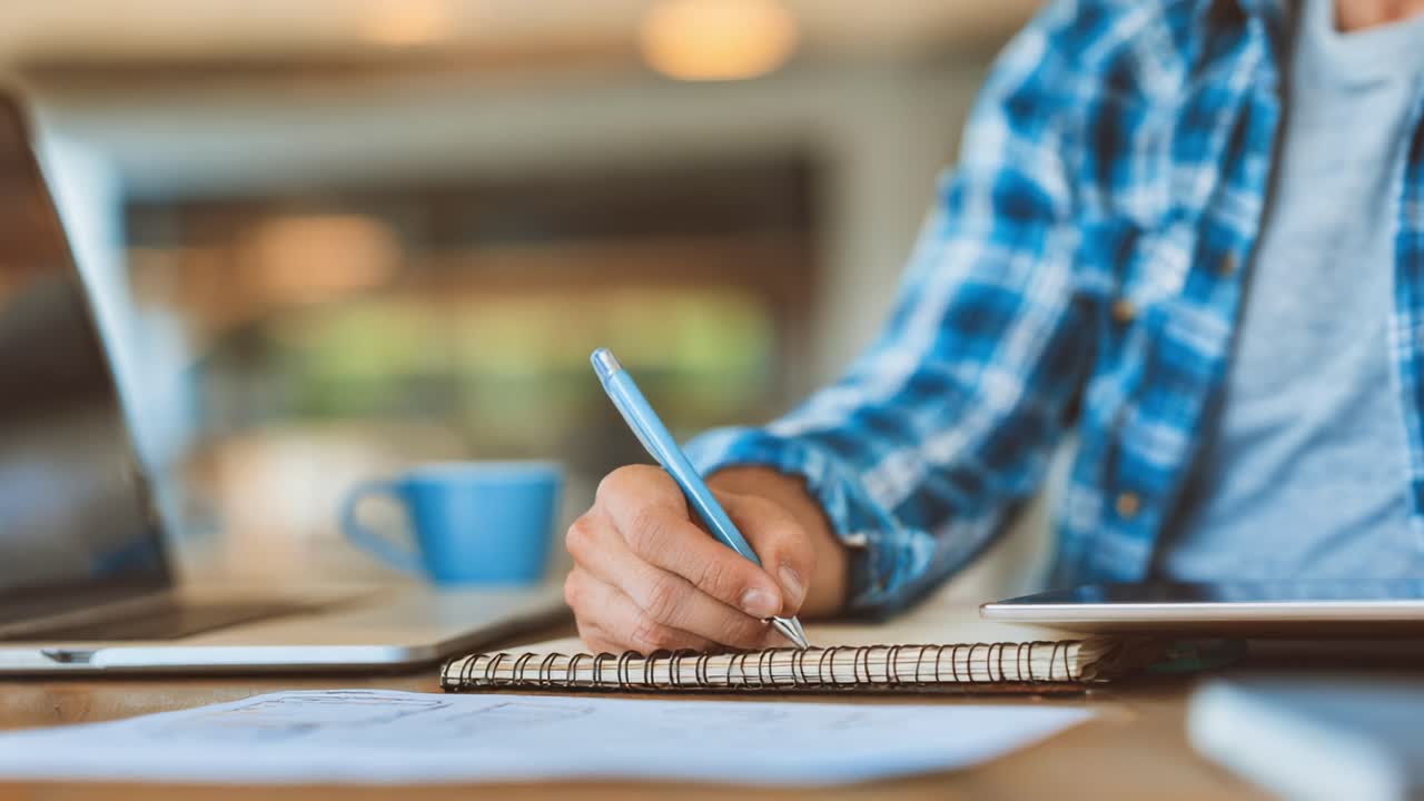 A Focused Moment of Creativity: A Person Engaged in Writing Notes or Ideas in a Notepad, Surrounded by Laptops and a Coffee Cup in a Bright Workspace Setting