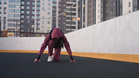 active woman kneeling in starting sprint position on curved sports court asphalt next to white barrier topped with fence in urban environment wearing maroon hoodie black vest and white running shoes