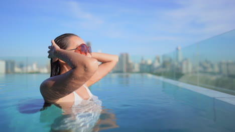 Joven-Asiática-Parada-Medio-Sumergida-En-La-Piscina-En-Una-Azotea,-Levantando-Los-Brazos-Y-Tocando-Su-Largo-Cabello-Húmedo-Con-Un-Paisaje-Urbano-Borroso-En-El-Fondo