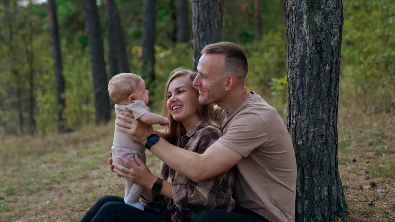 Caucasian parents sit in the woods holding their newborn child. Happy couple talk, smile and kiss their lovely infant.