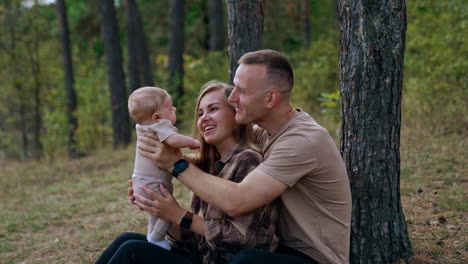 caucasian parents sit in the woods holding their newborn child. happy couple talk, smile and kiss their lovely infant.