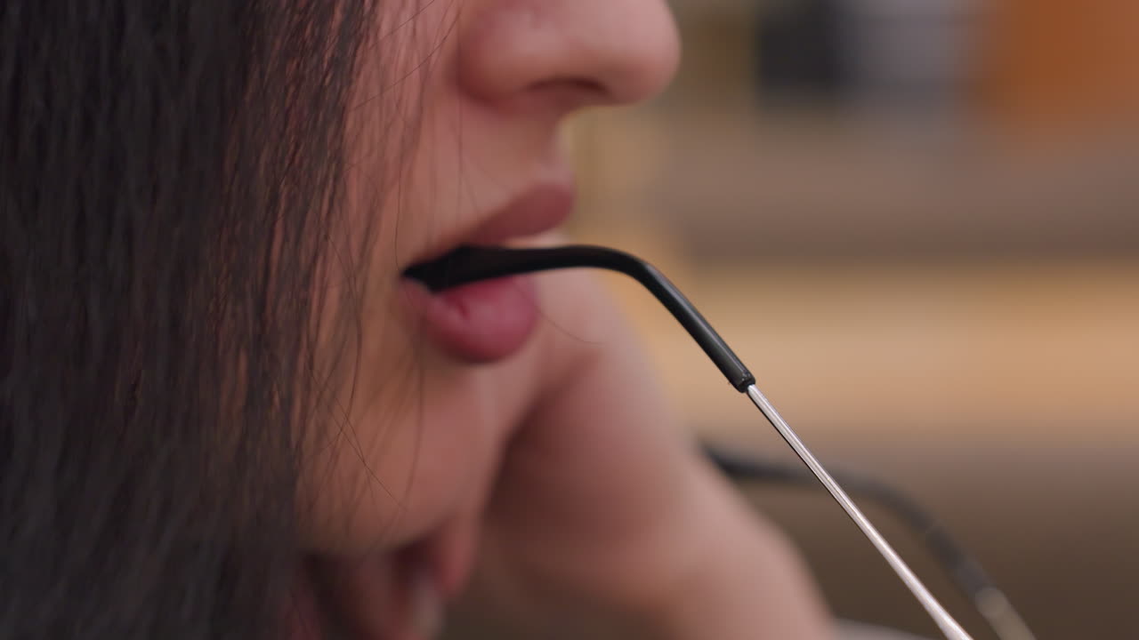 Close up of fair skinned woman holding eyeglasses with lips in focus as she gently grips frame with mouth, creating intimate, stylish detail in soft indoor setting with shallow depth of field