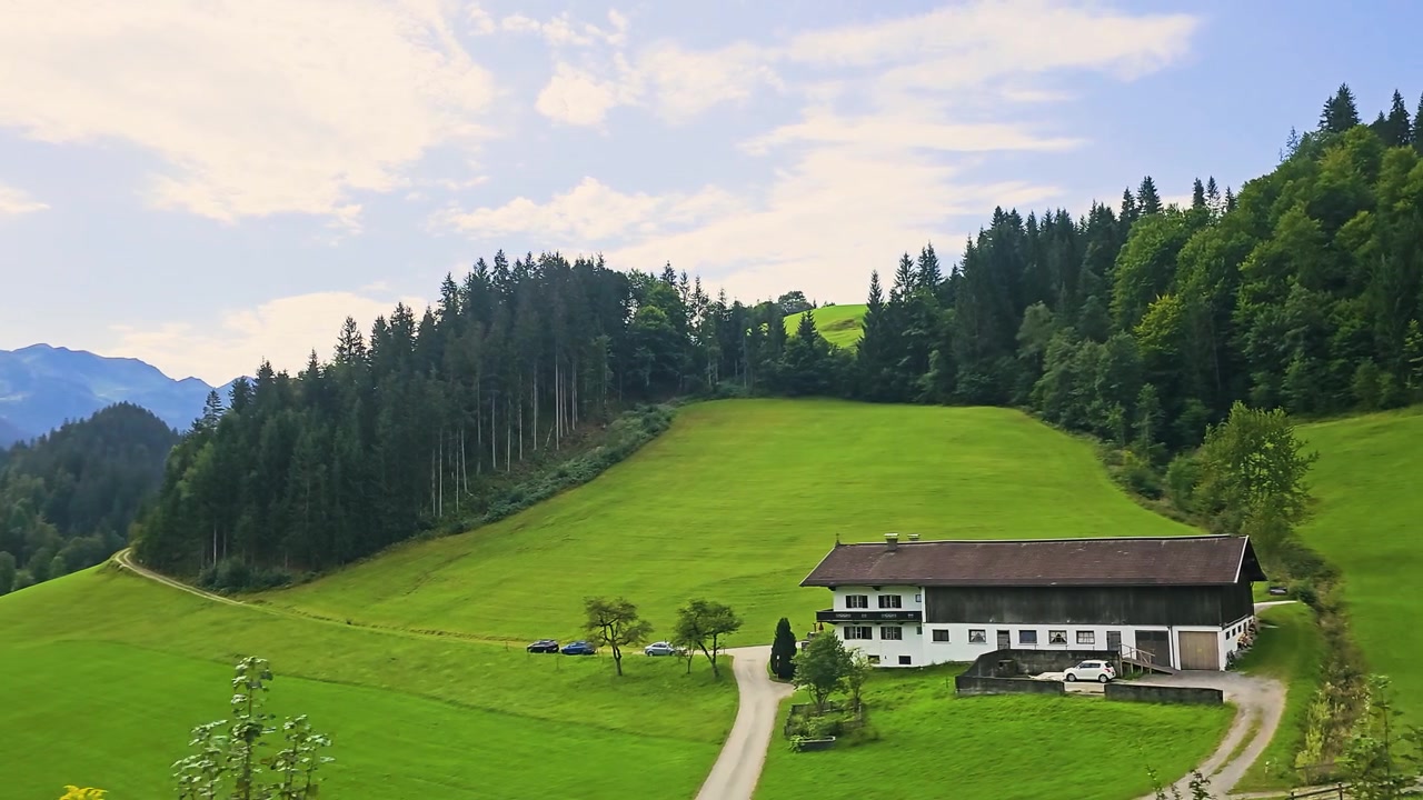 Paesaggio alpino da un treno, con prati soleggiati, chalet, colline boscose e vette montane che scorrono in una ripresa fluida di viaggio attraverso una regione montuosa.